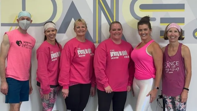 A group of one mand and five women, all wearing pink shirts, stand in front of a wall that says “dance” after completing the Pink Out Jazzercise event.